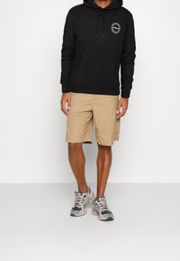 Zwarte hoodie met logo op de borst, gecombineerd met beige shorts. Model draagt grijs en zwarte sportchoenen met paarse accenten.