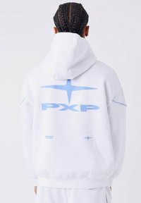 Persoon met gevlochten haar, die een witte hoodie draagt met een blauw "PXP"-logo en ster-symbolen op de achterkant, met de rug naar de camera gekeerd.