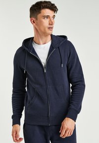 Marineblaue Kapuzenjacke mit Reißverschluss, Kordelzugkapuze, Fronttaschen, gerippten Bündchen und einer weichen Textur, getragen über einem weißen Shirt.