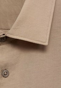 Beige puuvillapaita, jonka kauluksessa on näkyvä nappi ja siisti ompeluyksityiskohta sileällä kankaan pinnalla.