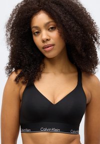 Crna braletica s V-izrezom, izrađena od glatkog materijala. Ima brendirani elastični pojas ukrašen bijelim natpisom.