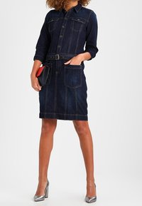 Jeansskjolklänning i mörkblått, med knappstängning, två bröstfickor, bälte i midjan och rak skärning. Matchas med silverfärgade högklackade skor.