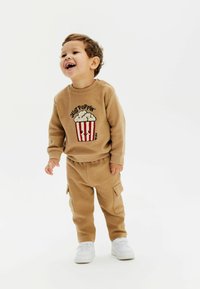 Tan sweater met popcornontwerp en rode strepen, gecombineerd met bijpassende tan broek met zakken. Witte sneakers maken het outfit compleet.