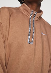 Brun pullover i mjukt tyg, med halvmatchning och inramning i hundtandmönster samt en liten vit logotyp på bröstet.