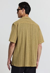 Uomo di spalle che indossa occhiali da sole neri, una maglietta a maniche corte beige sfumata e pantaloni neri, su uno sfondo chiaro e semplice.