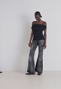 Nuoga pečių juoda palaidinė, derinama su nusidėvėjusiais pilkais pločiais džinsais ir juodomis šlepetėmis su apvaliais vilkės taškeliais, prieš šviesią foną.