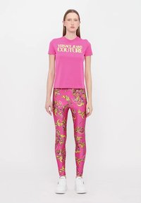 Roza bombažna majica s tekstom "VERSACE JEANS COUTURE" in roza legice z rumenim cvetličnim vzorcem, v kombinaciji z belimi supergami.