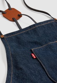 Delantal de mezclilla con tela azul oscura y costuras doradas en contraste, un bolsillo frontal y un detalle de hexágono de cuero marrón en el cuello.
