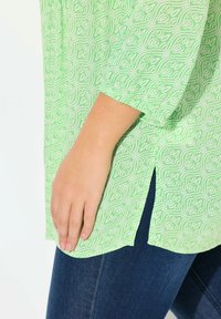 Haut vert à motifs avec de longues manches et fentes latérales, porté sur un jean bleu foncé. Le tissu présente un motif en vagues blanches.