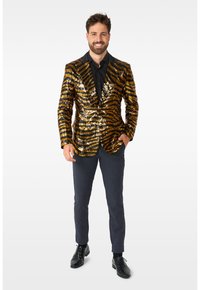 Gold- und schwarz gepunkteter Blazer mit tailliertem Schnitt, kombiniert mit einem schwarzen Hemd und dunklen Hosen. Schwarze Schuhe runden das Outfit ab.
