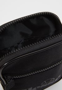 Czarna torba z tkaniny o gładkiej fakturze, zapinana na zamek błyskawiczny, z detalem logo. Wnętrze wyłożone miękkim materiałem.