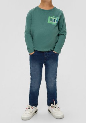 Camisa verde de manga larga con el gráfico "SIEMPRE VUELA MÁS ALTO", combinada con jeans de mezclilla azul y zapatillas blancas con detalles en negro.