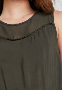 Ärmlös, olivgrön topp i lättviktsmaterial med en genomskinlig panel och dekorativ sömnad längs halsringningen. Slät textur.