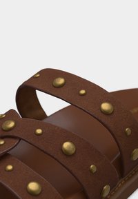 Sangles de sandales en cuir marron avec des clous ronds en laiton de différentes tailles, montrées en gros plan sur un fond blanc.