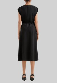 Eine Frau steht mit sichtbarem Rücken da, trägt ein ärmelloses schwarzes Midikleid und schwarze High Heels vor einem schlichten grauen Hintergrund.