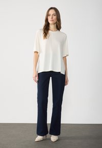 Hvit kortermet topp med rund hals, pairt med mørkeblå utsvevende jeans og beige teksturerte høye sko på et grått gulv.