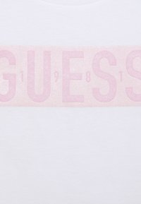 Bílý tričko s velkým růžovým texturovaným potiskem se slovem "GUESS" a číslicemi, umístěným na hrudi. Bavlněný materiál.