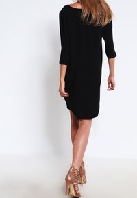 Svart löst sittande klänning med trekvarts ärmar, något rundad nederkant, i kombination med ljusbruna sandaler med hög klack. Slät textur.