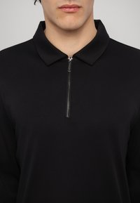 Svart, langermet polo-skjorte med krage og halv-zip foran. Glatt stofftekstur, minimalistisk design uten mønstre eller utsmykninger.