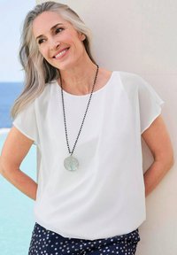 Weiße, lockere Bluse mit kurzen Ärmeln, kombiniert mit navyblauen Punkten-Hosen. Langer schwarzer Halskette mit einem runden Anhänger.