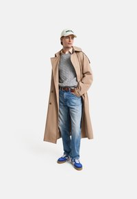 Beige trenchcoat, grå kabelstriketrøje, lyseblå jeans og blå sneakers med hvide og brune accenter. Hvid kasket med grøn tekst.