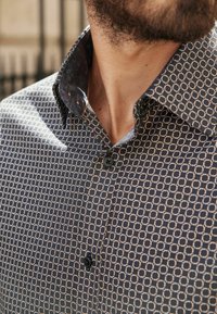 Chemise bleu marine avec un motif géométrique de cercles entrelacés en beige. Présente un col floral contrastant et un seul bouton noir.