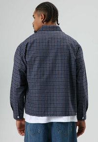 Marineblauw geruit overhemd met lange mouwen, met blauwe en rode ruitpatronen, manchetten met knopen en een licht losse pasvorm. Draagt over een wit T-shirt.