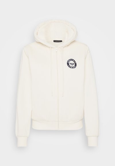Bijela zip-up dukserica s teksturiranom unutrašnjošću, sadrži okrugli mornarsko plavi logo na lijevoj strani grudi, elastične manžetne i pojas.