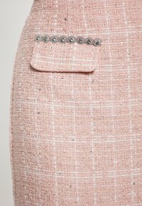 Il tessuto tweed rosa presenta una superficie tessuta e texturizzata con accenti bianchi. Include una piccola tasca con decorazioni in tonalità argentata.