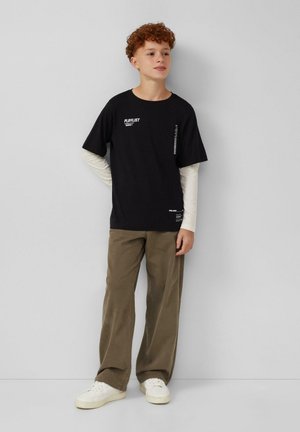 Un adolescent aux cheveux roux bouclés se tient contre un mur, portant des manches longues noires et blanches superposées, un pantalon marron et des baskets blanches.