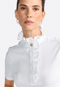 Polo shirt bianco a maniche corte con colletto arricciato e patta, dotato di un bottone decorativo all' scollo. Texture in morbido cotone.
