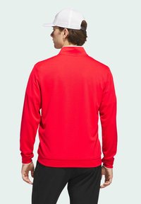 Long-sleeve red polo shirt with a collar, made of textured fabric. Paired with black pants. Worn with a white perforated cap.