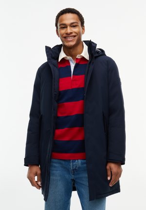 Giovane uomo sorridente, indossa un cappotto con cappuccio blu navy sopra una maglia da rugby a righe rosse e blu navy e jeans blu, in piedi davanti a uno sfondo semplice.