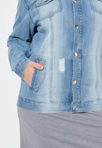 Vaaleansininen farkkutakki, jossa on hopeiset napit, kaksi rinnapiliä ja kulunut alue. Sileä malli ja hieman haalistunut rakenne.