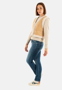 Gilet en suède tan avec doublure en shearling crème, superposé à un pull en maille beige. Jean bleu slim et baskets blanches avec accents bleus et oranges.
