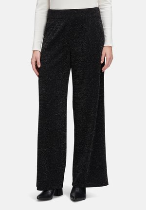 Vrouw in zwarte glitterachtige wijde broek, witte lange mouwen top en zwarte enkellaarsjes, staand op een witte achtergrond.