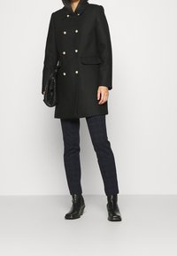 Abrigo negro de doble botonadura con botones dorados, mangas largas y bolsillos laterales, combinado con pantalones oscuros de cuadros y botas negras hasta el tobillo.