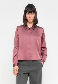 Camicia di raso mauve con colletto a punta, maniche lunghe e chiusura con bottoni; abbinata a pantaloni grigi su misura. Texture liscia, design cropped.