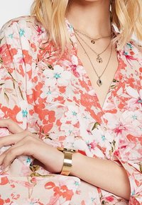 Haut cache-coeur floral aux nuances roses et blanches, avec des accents bleus. Colliers en or superposés et montre en or texturée au poignet.