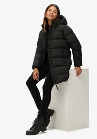 Chaqueta acolchada negra con capucha, que presenta un cierre de cremallera en la parte delantera, bolsillos laterales y un diseño elegante, llevada sobre un conjunto negro y botas negras.