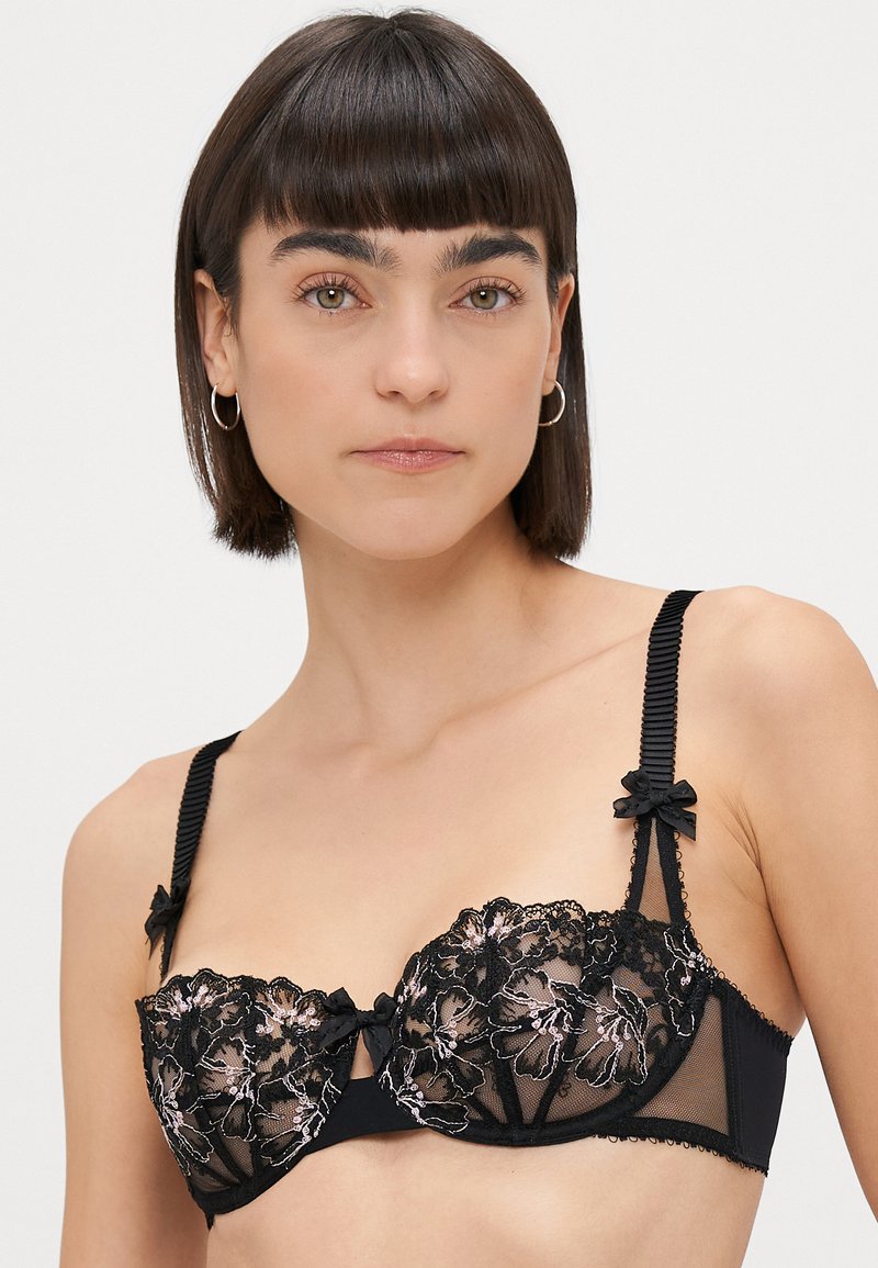 Reggiseno nero con dettagli in pizzo floreale, supporto con stecche, e fiocchi in satin nero sulle spalline regolabili. Coppe in rete trasparente e tessuto liscio.