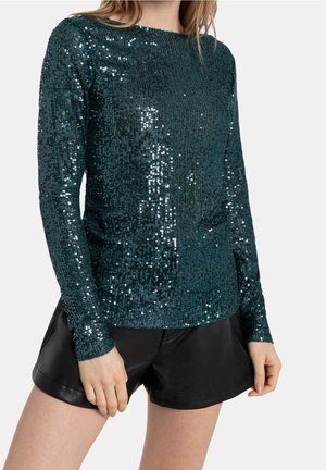 Maglietta a maniche lunghe di color teal, coperta di paillettes lucide; design aderente con scollatura rotonda; abbinata a pantaloni corti di pelle nera.