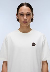 Tricou alb din bumbac, cu o emblemă rotundă de culoare neagră și portocalie pe piept. Potrivire lejeră și mâneci scurte. Textură netedă.