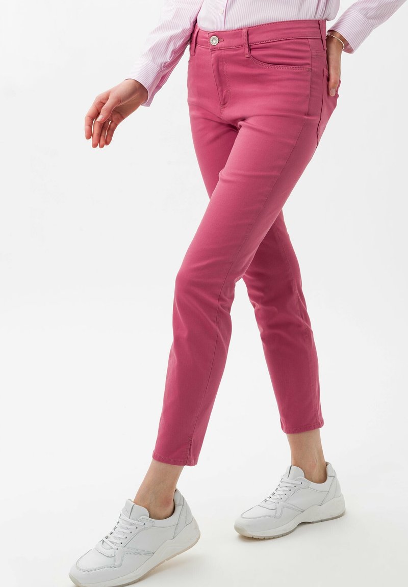 Femme portant un pantalon slim rose et des baskets blanches, debout avec une main dans la poche et l'autre bras détendu.