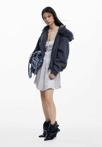 Hoodie oversize bleu foncé avec col doublé de fourrure, associé à une robe gris clair. Sac gris transparent avec des accents bleus, et bottes noires décontractées.