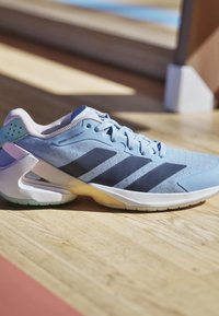 Hellblauer Sportschuh mit schwarzen Streifen und weißer Sohle auf Holzboden mit Sonnenlicht und Schatten.