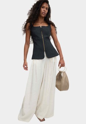 Kvinde med langt krøllet hår iført sort ærmeløs top med lynlås, brede hvide bukser, holder beige håndtaske med guldringe som håndtag.