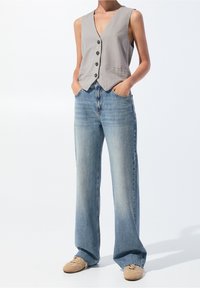 Chaleco gris sin mangas con escote en V, que cuenta con cinco botones y bolsillos, combinado con unos pantalones vaqueros de pierna ancha en azul claro y zapatos beige.