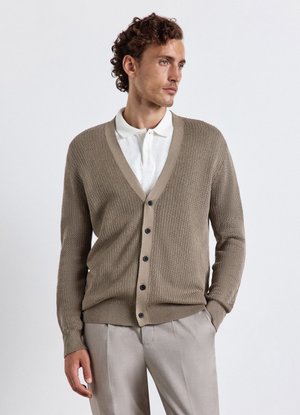 Uomo che indossa un cardigan marrone chiaro abbottonato sopra una polo bianca con pantaloni beige, guarda verso destra su uno sfondo semplice.