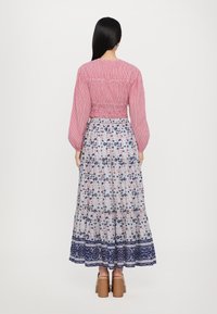 Langes Kleid mit einem roten, gerafften Oberteil, langen Ärmeln und einem gemusterten, blauen Blumenrock. Mit gestuften Rüschen am Saum. Braune Blockabsatzschuhe.